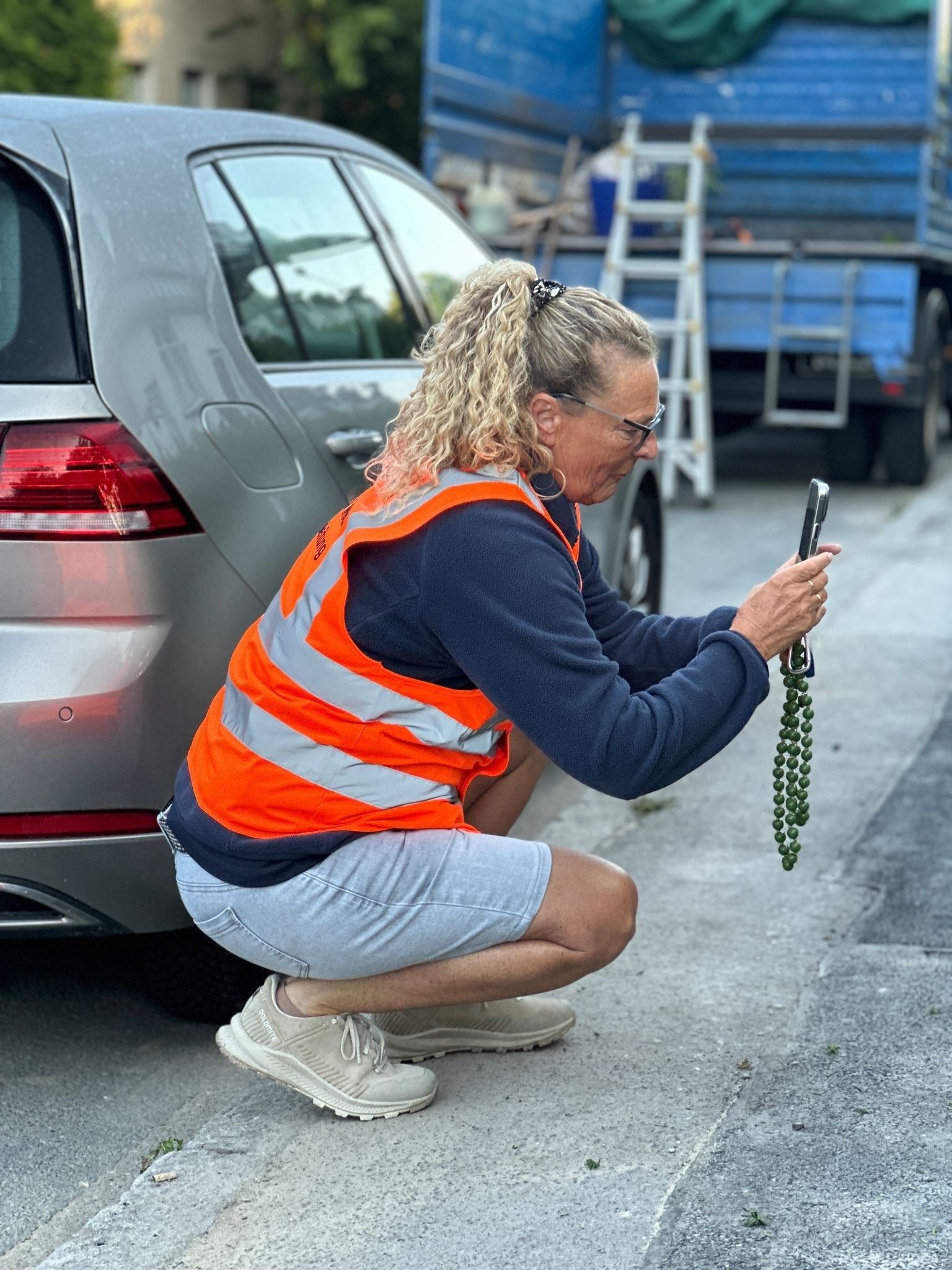 Eine Frau in einer Sicherheitsweste hockt und untersucht einen Gegenstand auf dem Boden neben einem Auto.