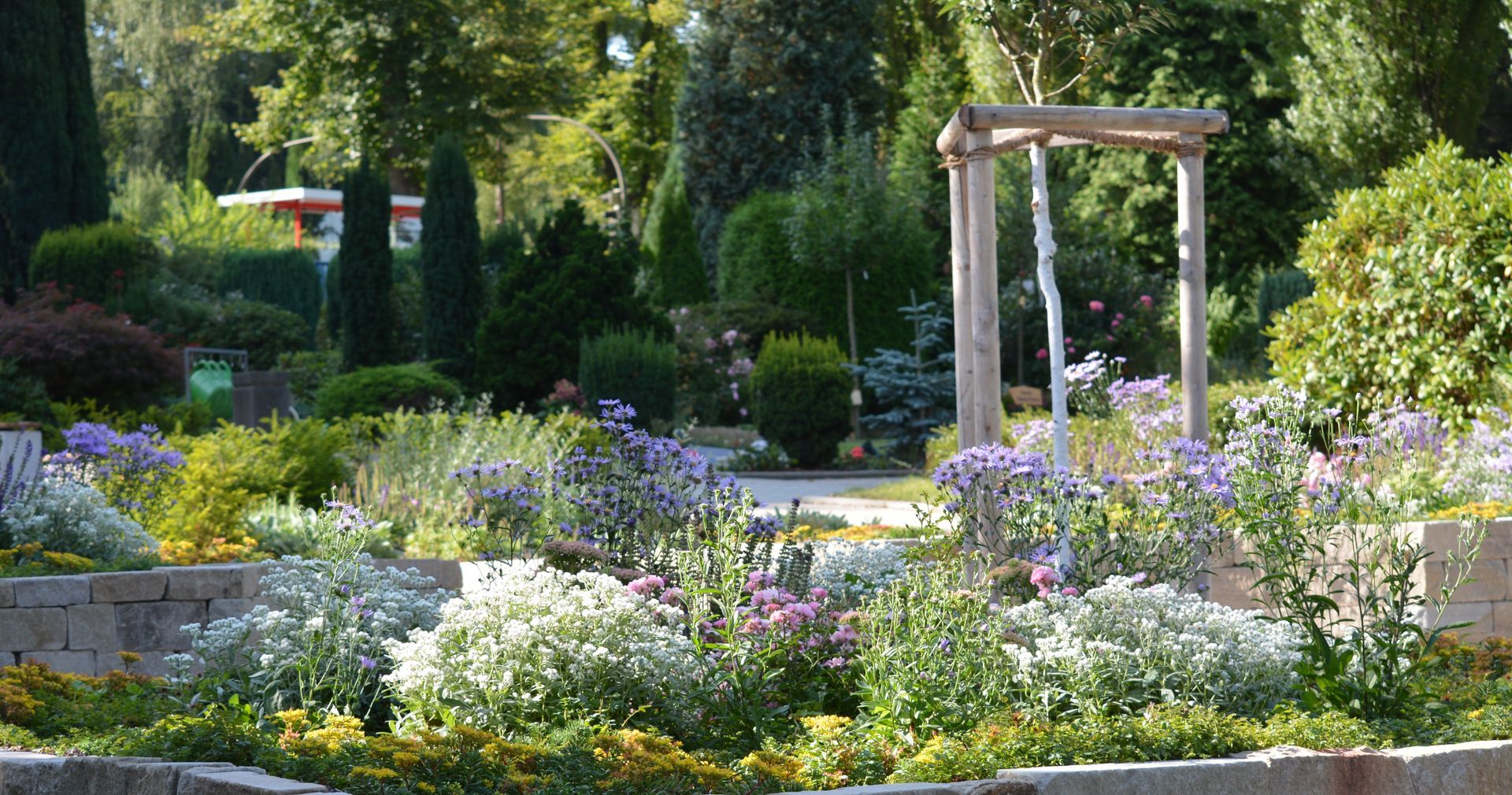 Ein von Steinmauern umgebener, terrassenförmig angelegter Garten mit verschiedenen Pflanzen und Blumen, darunter eine Holzschaukel und üppige Bäume unter sonnigem Himmel.