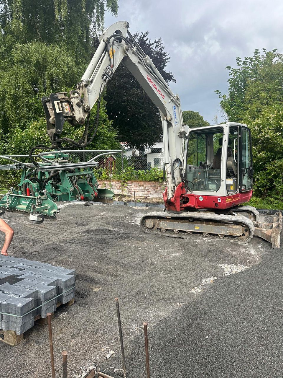 Ein Bagger arbeitet auf einer Kiesfläche neben Grünflächen. Im Vordergrund ist eine Palette mit Pflastersteinen zu sehen.