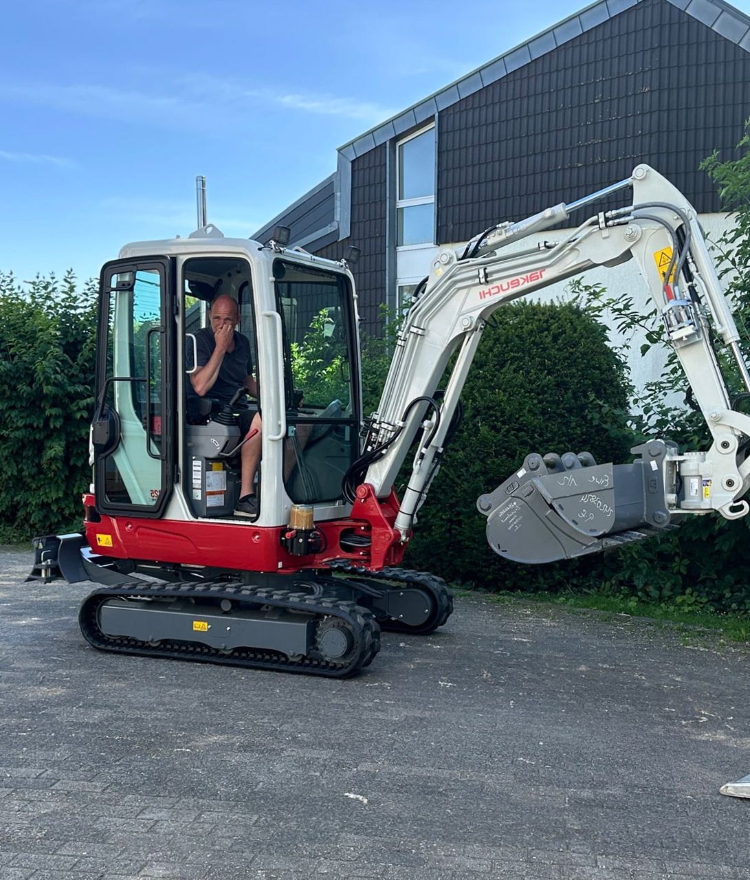Ein Mann bedient vor einem Gebäude einen weiß-roten Minibagger mit Schaufelaufsatz.