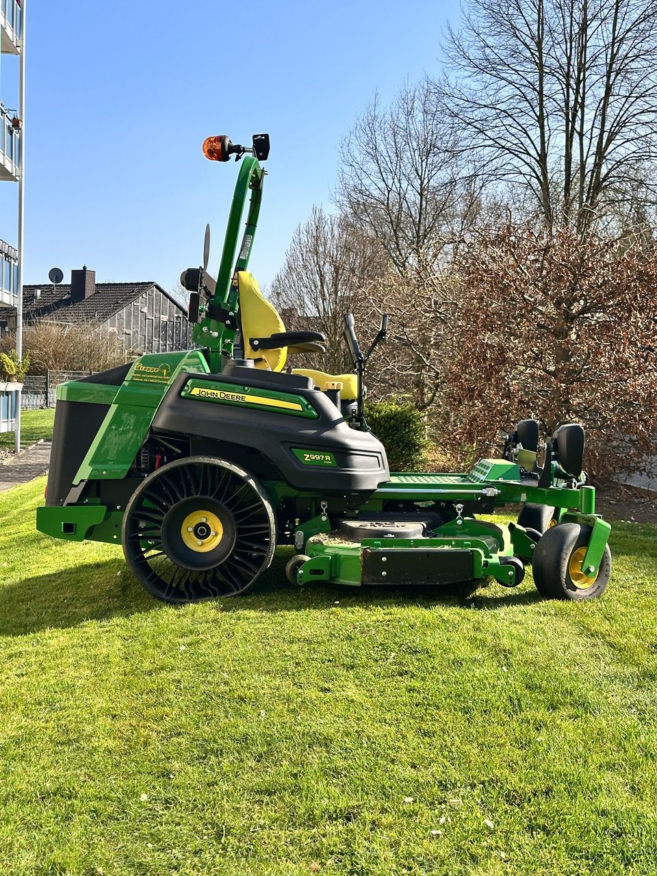 Ein John Deere Zero-Turn-Rasenmäher auf einer Rasenfläche mit Gebäuden und Bäumen im Hintergrund. Farbgebung in Grün und Gelb.
