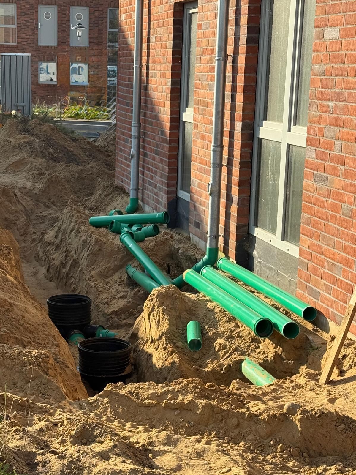 Baustelle mit in einem Graben neben einem Backsteingebäude verlegten grünen Rohren.