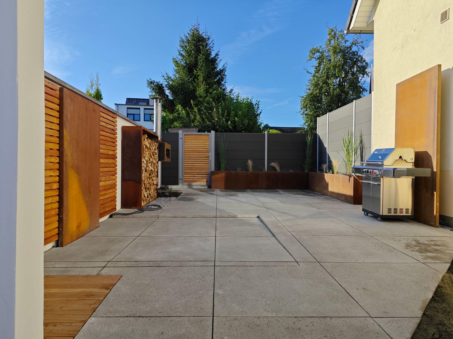 Terrasse mit Steinplatten, Metallgrill und Holzwänden unter blauem Himmel.