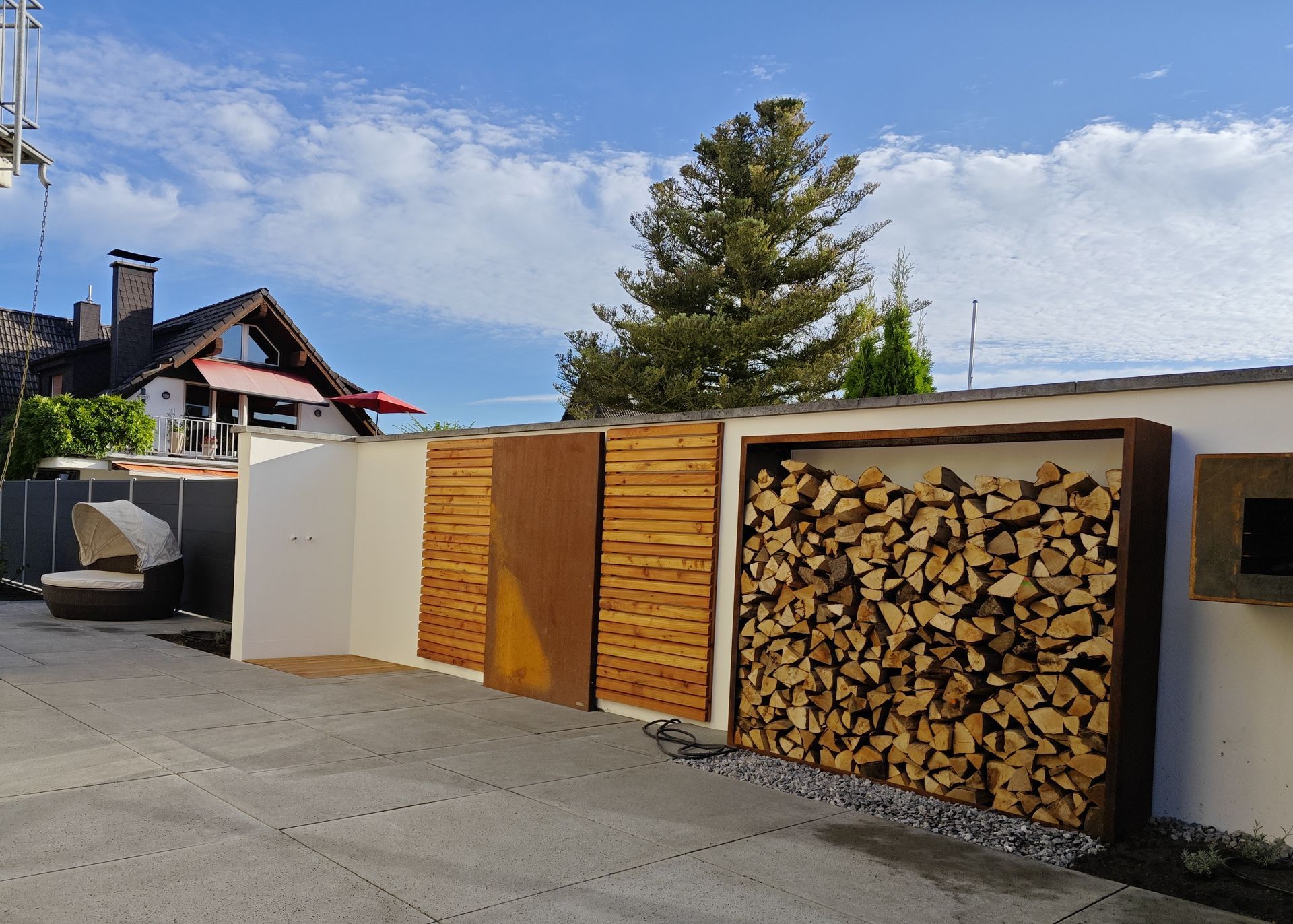 Eine Wand mit Holzregalen, Holzplanken und einer rostigen Metallplatte. Im Hintergrund heller Himmel.