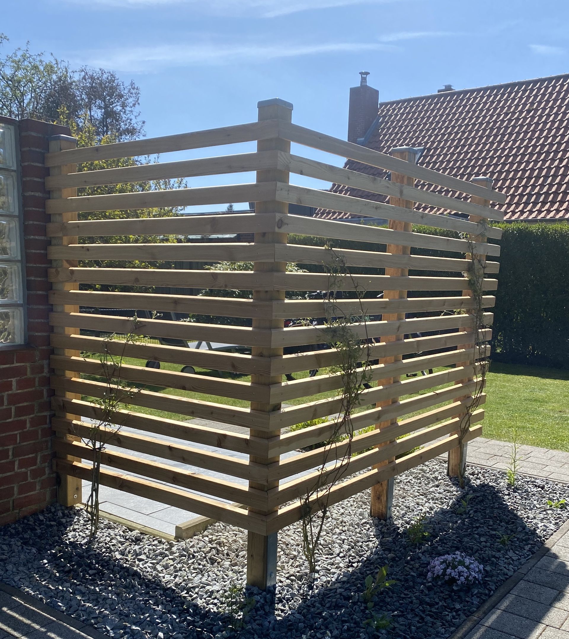 Eckförmiger Sichtschutz aus Holzlatten im Garten, auf Kies, mit Ziegelmauer.