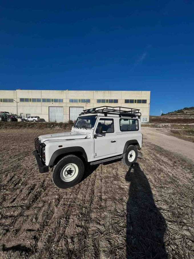 SUV Land Rover Defender de color blanco con portaequipajes en el techo sobre tierra bajo un cielo azul.