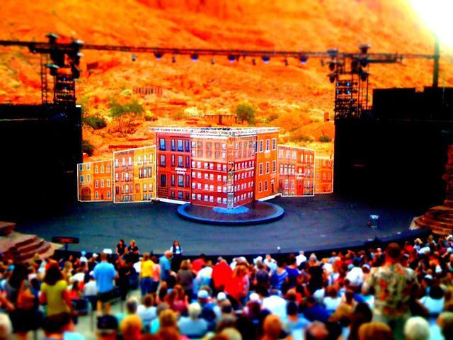 Crowd facing a miniature red building set on a stage against an orange hillside backdrop