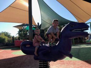 Two children on a purple dinosaur ride under shade sails at an outdoor playground