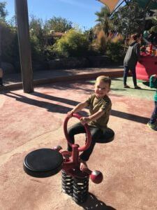 Child smiling on a playground balance toy under bright sun, with trees and other children nearby