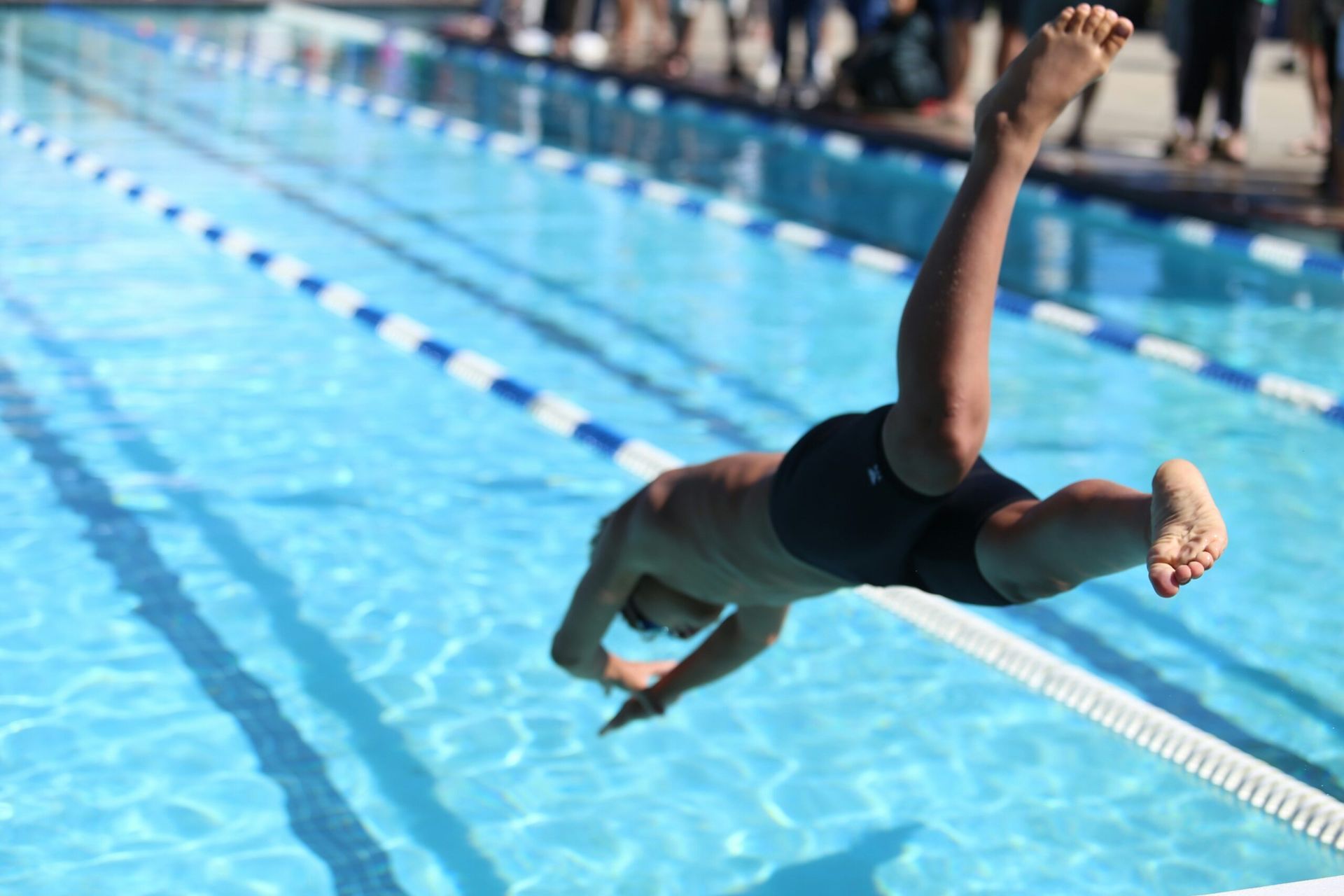 Swimmer diving into a bright blue pool, with spectators blurred along the edge