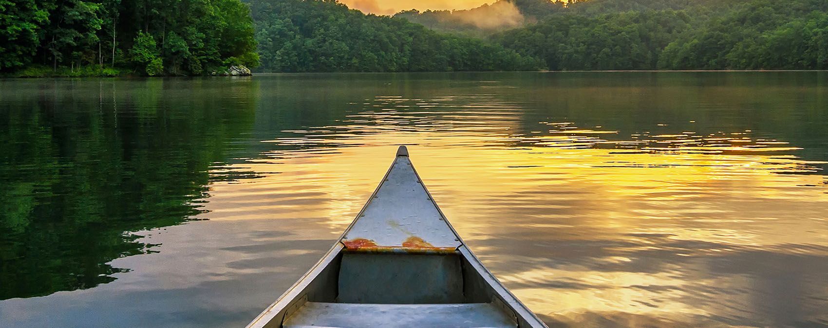 Canoe on a calm lake at sunset, reflecting golden light and surrounded by green forest and hills
