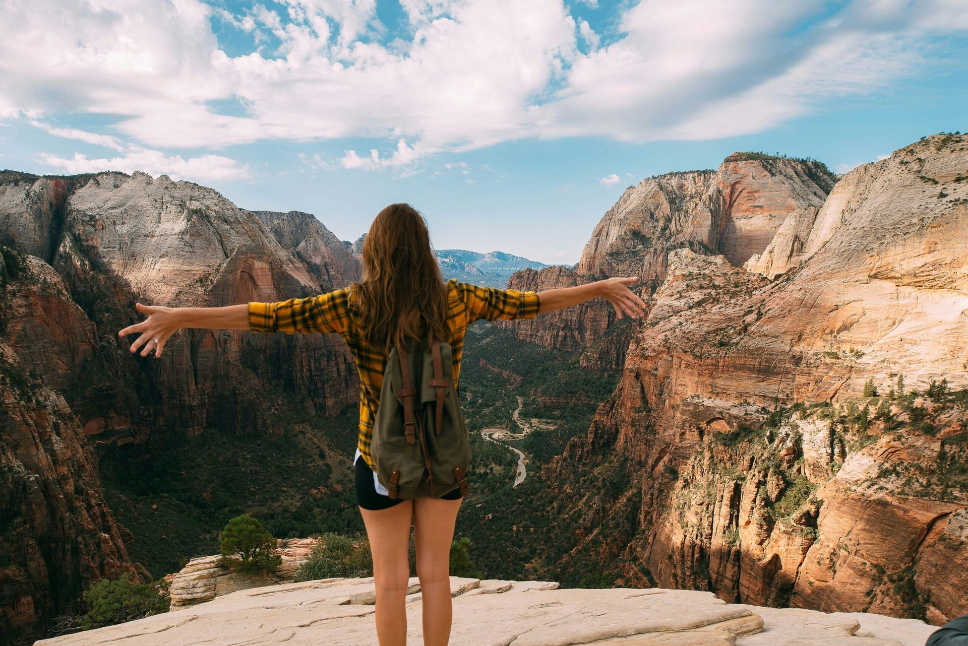 Person standing with arms outstretched on a cliff overlooking a canyon and mountains under a blue sky