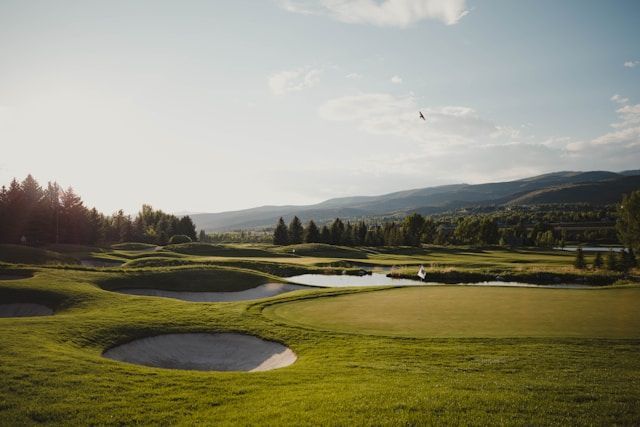 Sunlit golf course with sand bunkers, green fairways, trees, and distant hills under a clear sky