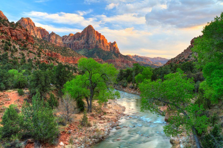 Rocky red cliffs over a green river valley under a bright, partly cloudy sky