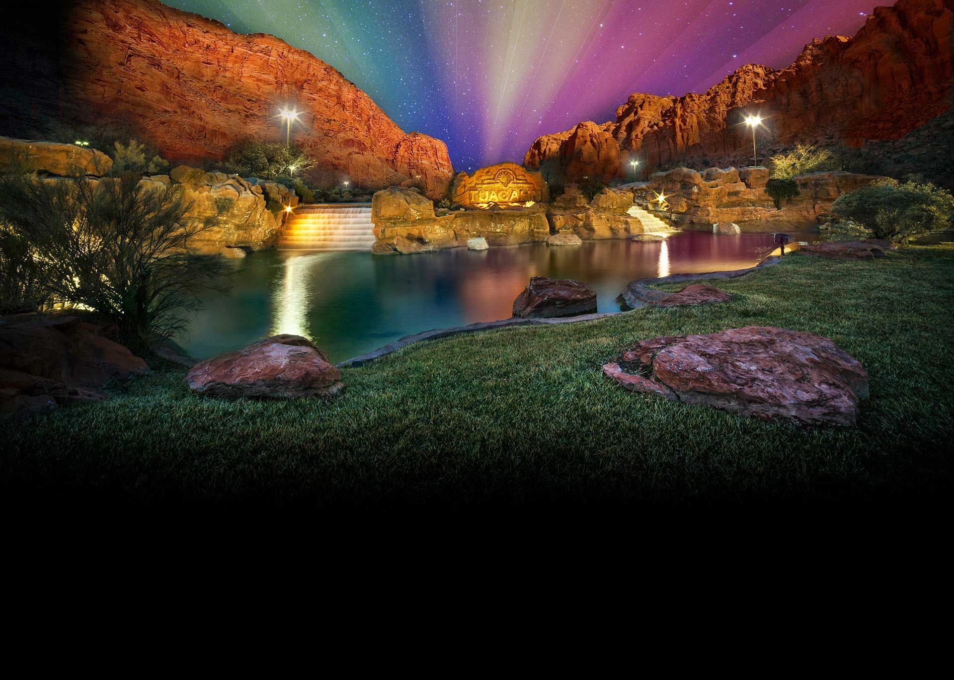 Nighttime canyon lake with glowing lights, orange cliffs, and colorful beams in the sky.