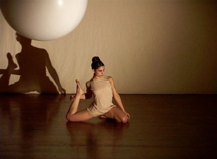 Bailarina con traje beige, arrodillada, con la espalda arqueada, buscando un pie. Gran esfera blanca