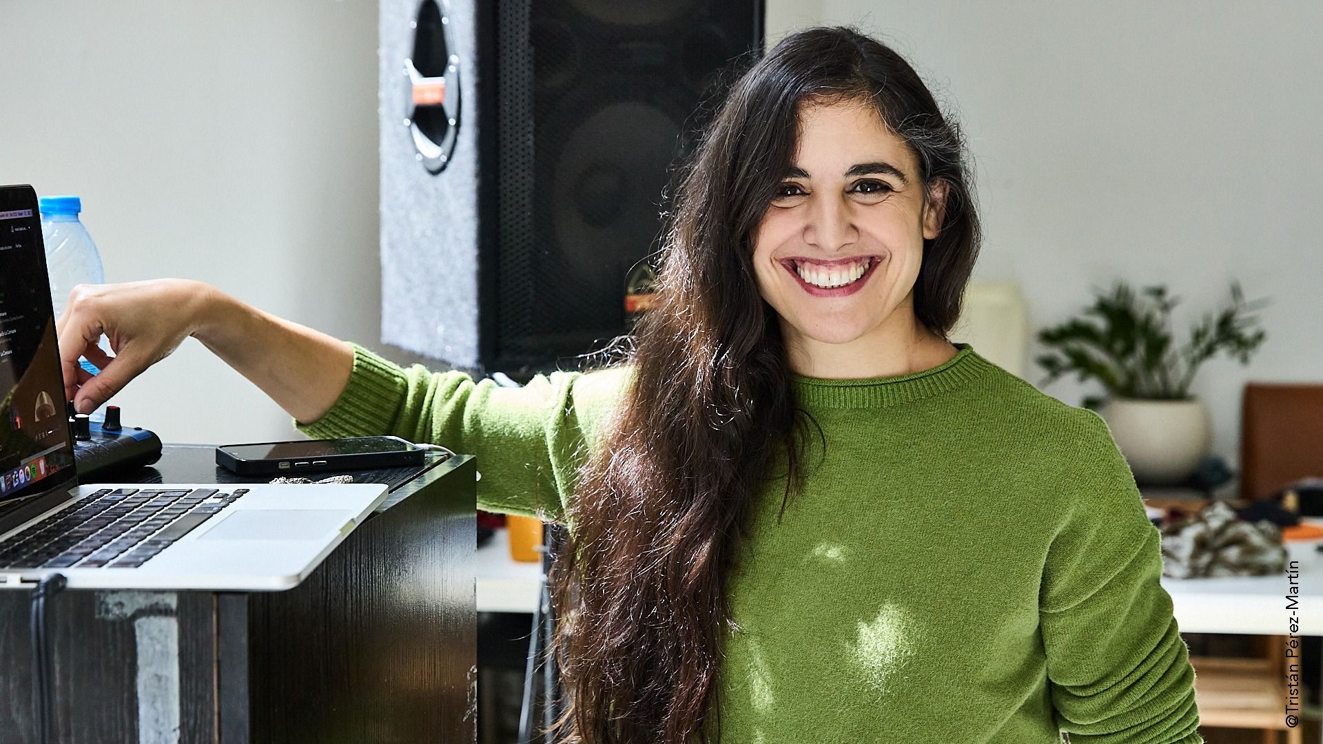 Mujer con cabello largo y oscuro sonriendo a la cámara, mirando hacia una computadora portátil; suéter verde; cerca de un alt