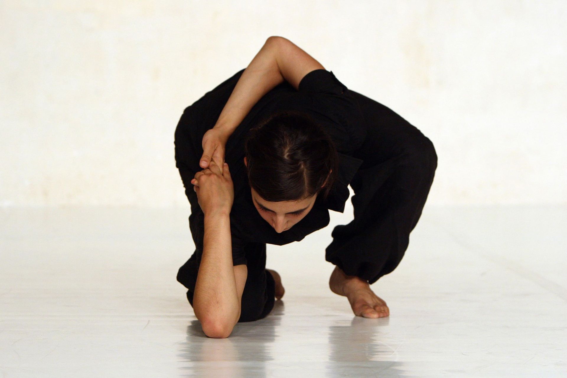 Persona vestida de negro contorsiona su cuerpo en el suelo, extendiendo los brazos detrás de la cabeza; posición neutral.