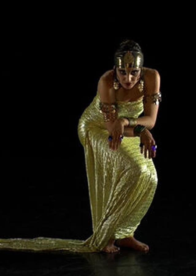 Mujer con vestido dorado, tocado y joyas en una pose dramática sobre un fondo negro.