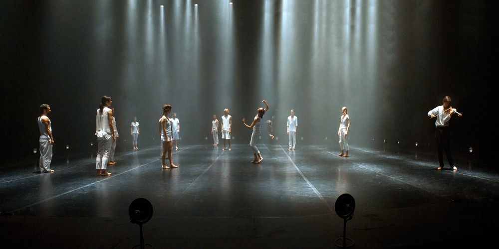 Bailarines vestidos de blanco actúan en un escenario iluminado por focos. Un bailarín está en el centro, en movimiento. Fondo