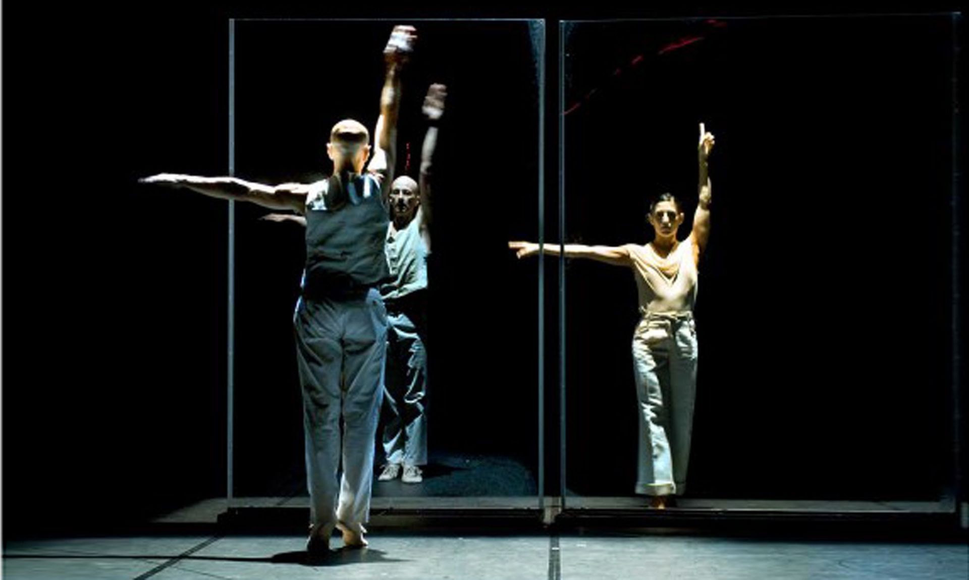 Tres bailarines en un escenario oscuro con paneles de vidrio; uno señala, dos tienen los brazos extendidos.