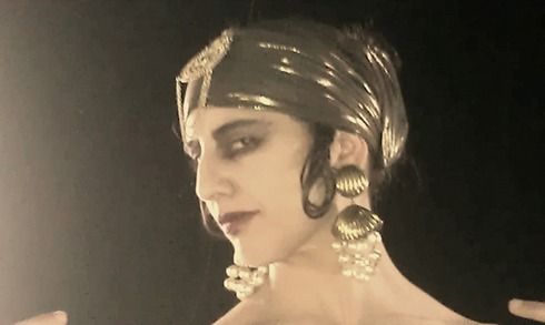 Mujer con diadema dorada vintage, pendientes, maquillaje oscuro, mirando por encima del hombro.