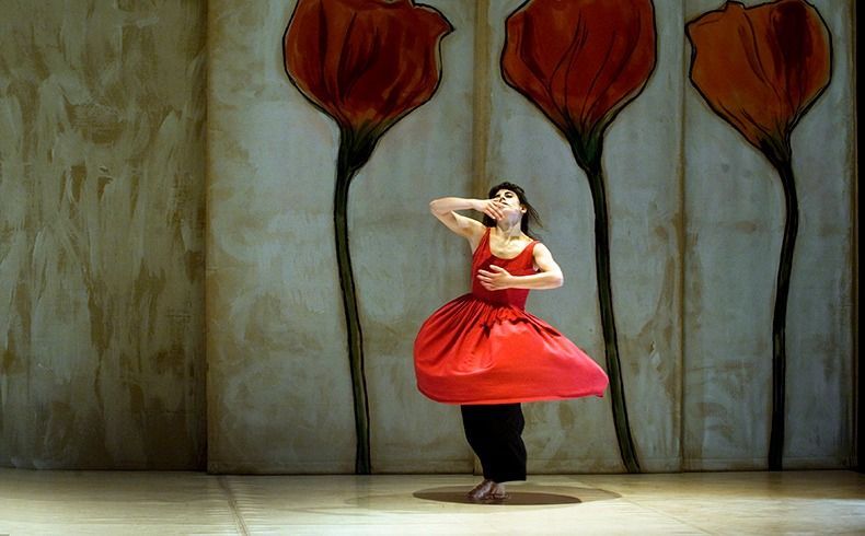 Bailarina con vestido rojo y pantalones negros, con los brazos en alto, se encuentra frente a grandes pinturas de flores