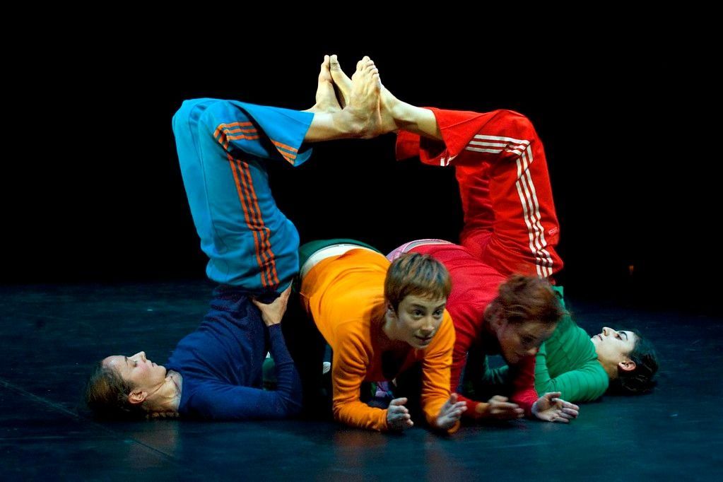 Bailarines con chándales coloridos forman un arco humano con los pies y posan sobre un escenario negro.