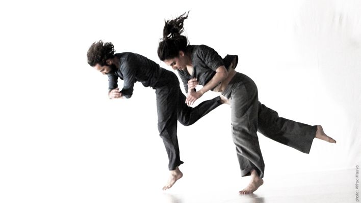 Dos personas vestidas de gris, en el aire, realizan un movimiento de artes marciales. Pies descalzos, fondo blanco.