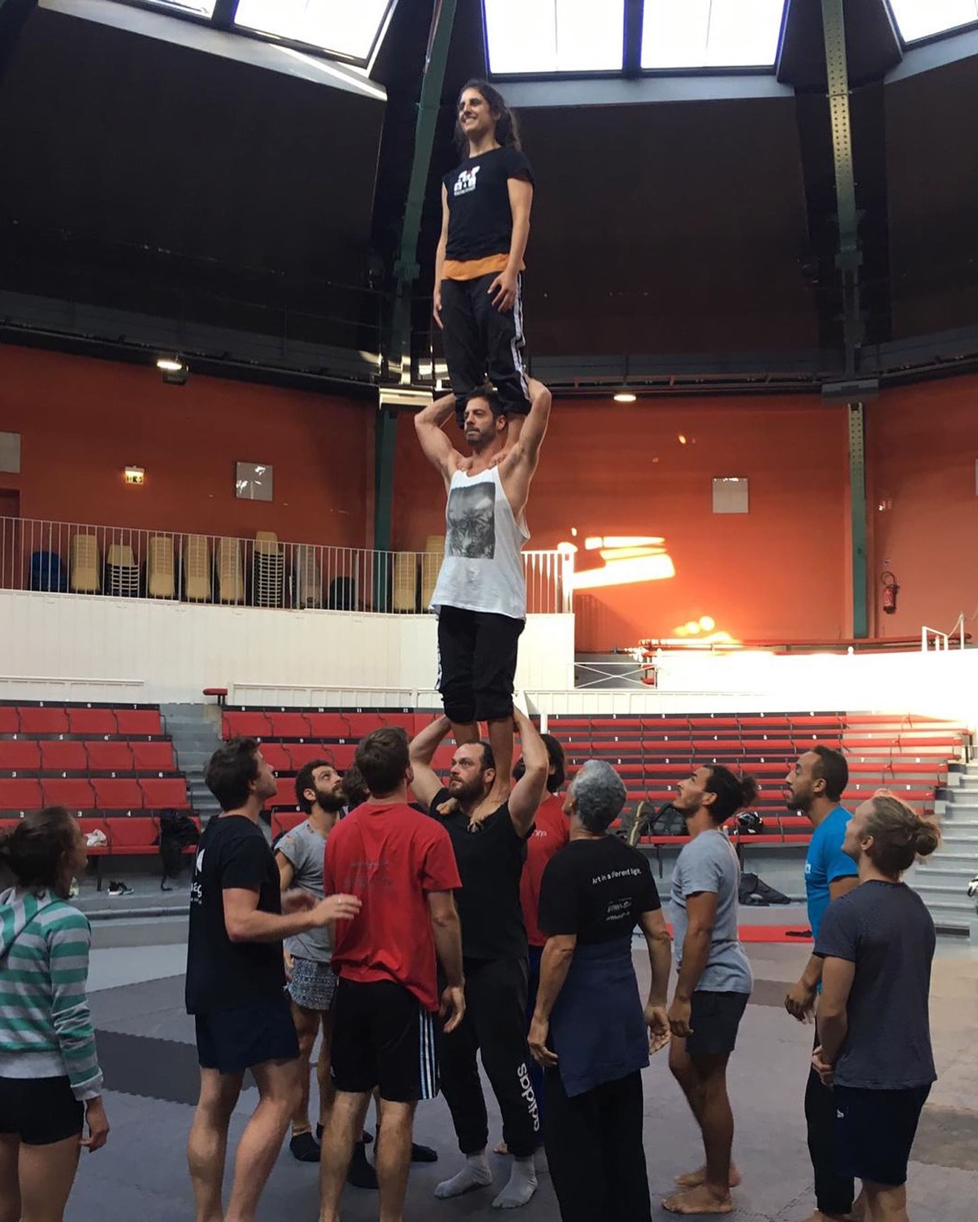 Acróbatas construyendo una pirámide humana en un gran estadio cubierto; una persona encima de otras dos, y más observando.