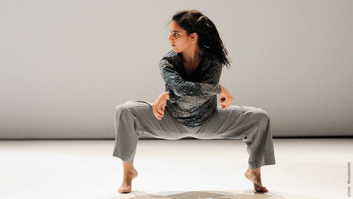 Bailarina con pantalón gris y camisa estampada en pose de sentadilla con los brazos cruzados, sobre un escenario luminoso.