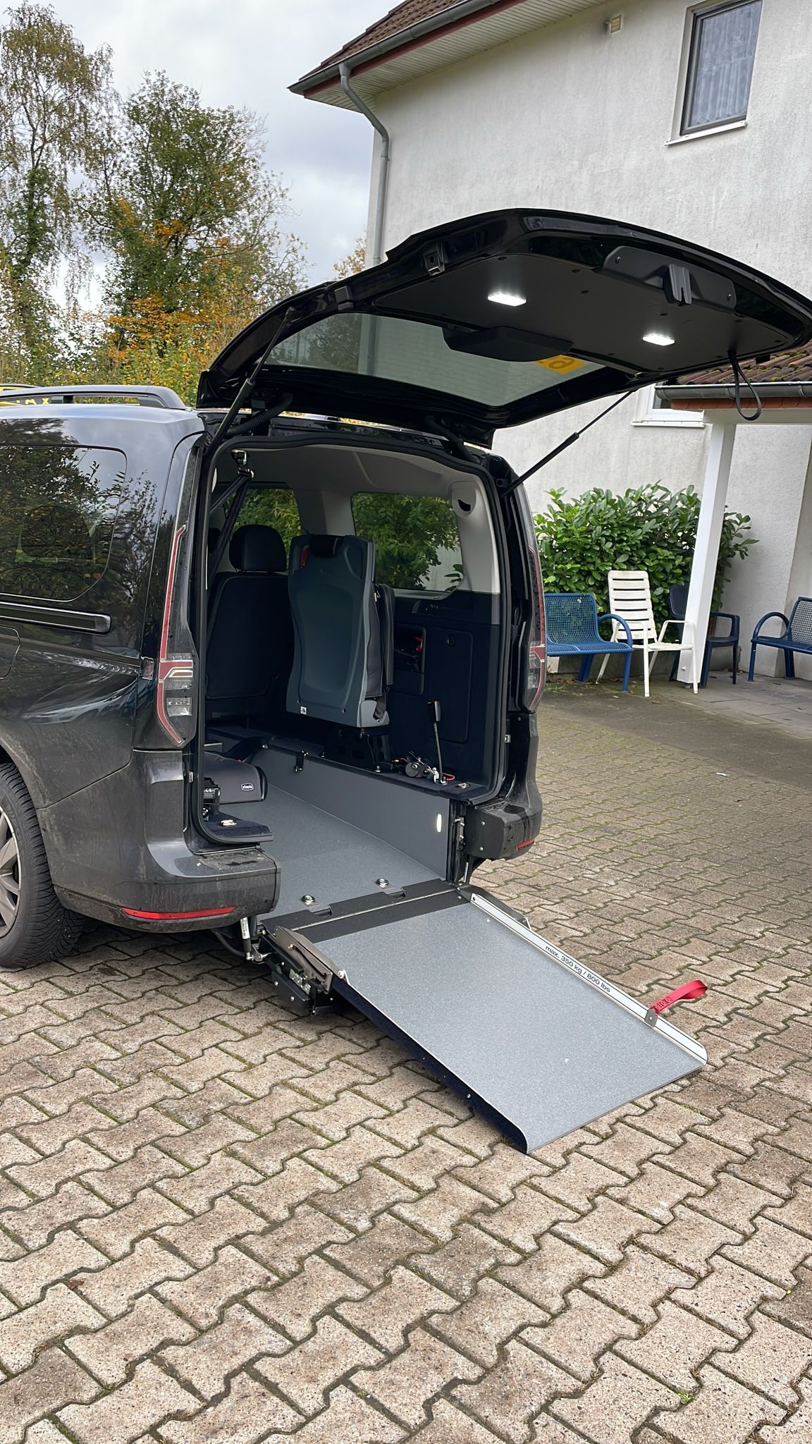 Ein schwarzer VW Caddy mit einer Rampe für den Rollstuhltransport ist auf einer gepflasterten Auffahrt geparkt.