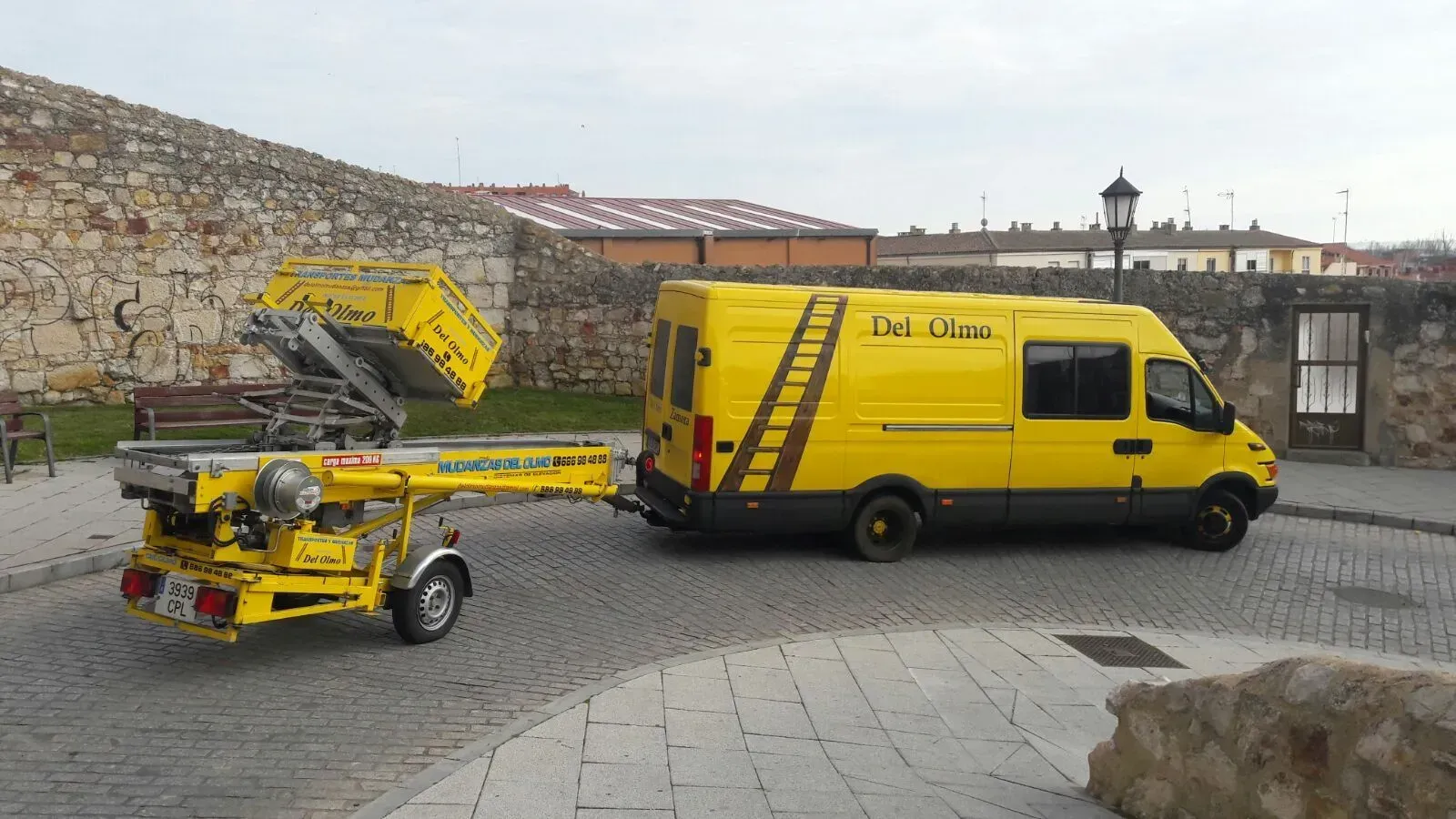 Furgoneta amarilla remolcando una plataforma elevadora amarilla sobre un remolque en una calle empedrada