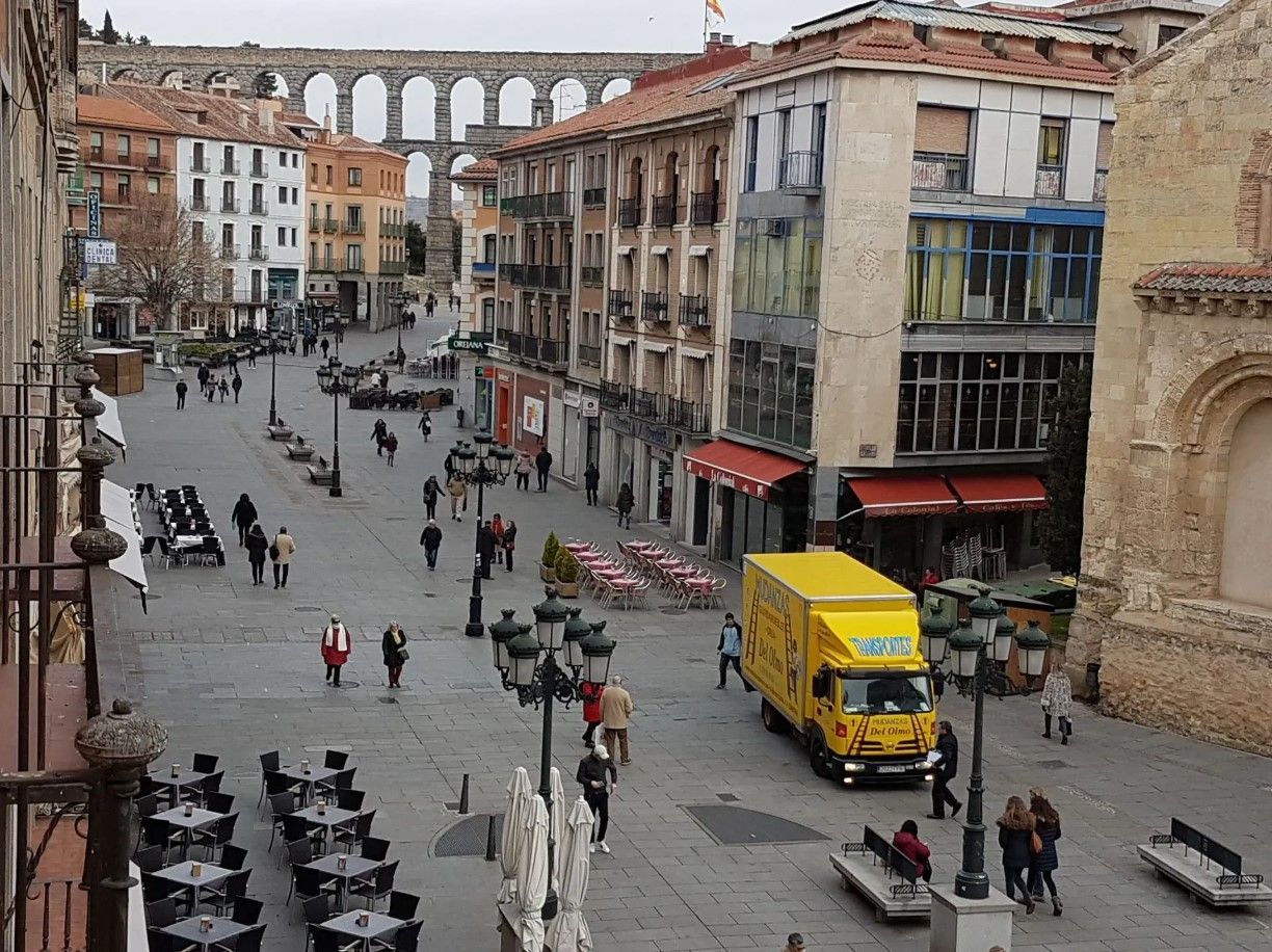 Escena callejera en Segovia, España, con el acueducto romano al fondo, gente caminando y restaurantes al aire libre.