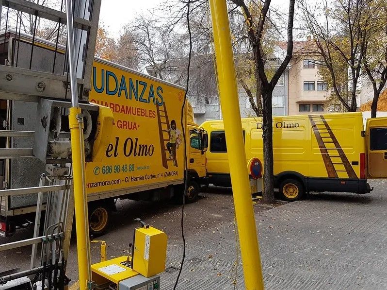 Camiones de mudanza amarillos estacionados en una calle con elevador y escalera; edificio al fondo.