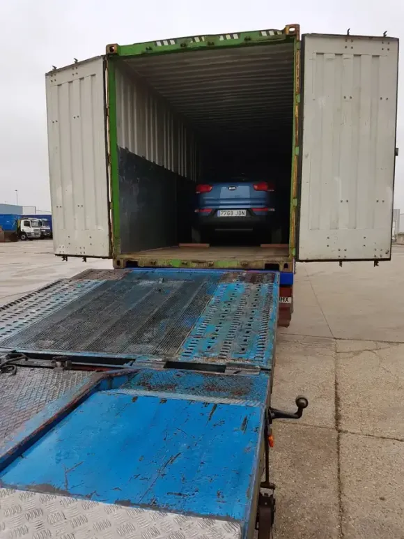 Coche azul dentro de un contenedor marítimo sobre una rampa azul. Puertas del contenedor abiertas; suelo de hormigón gris.