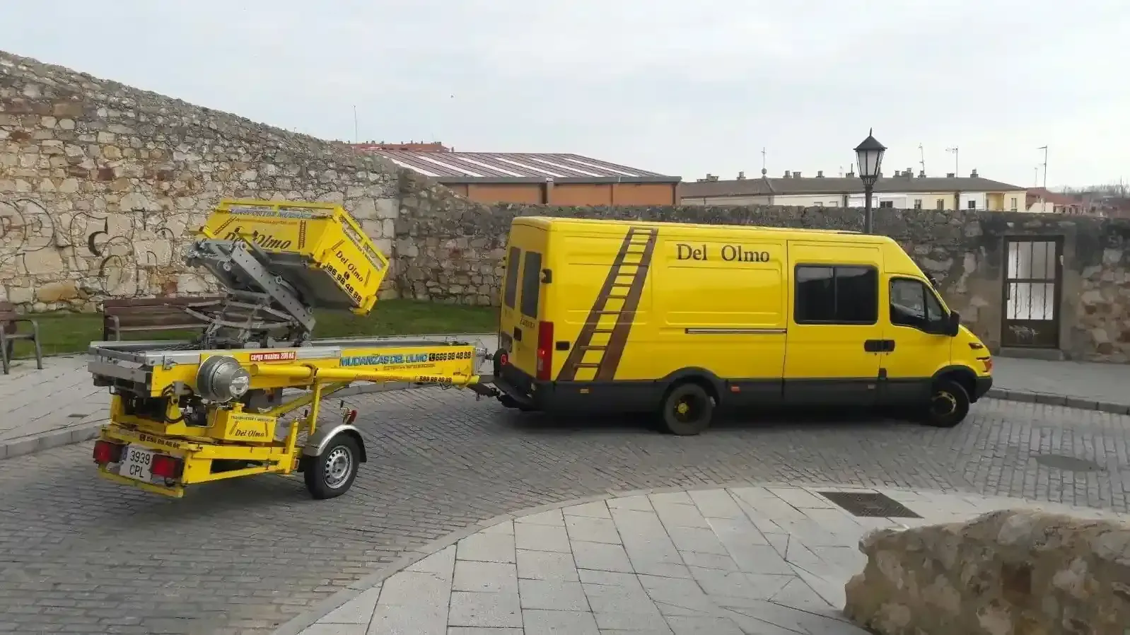 Furgoneta amarilla remolcando una plataforma elevadora amarilla en una calle adoquinada.
