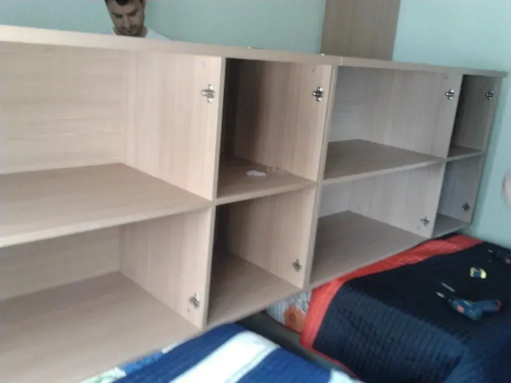 Un hombre monta un armario de madera de color claro con seis compartimentos abiertos; herramientas sobre la cama.