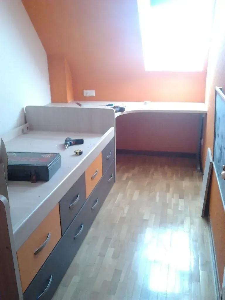 Dormitorio con cama y escritorio empotrados; cajones naranjas y grises; suelo de madera; techo inclinado con tragaluz.