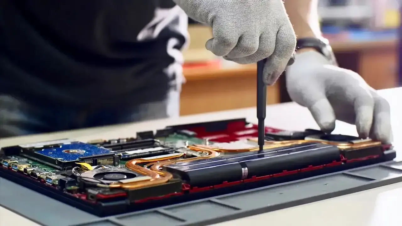Persona con guantes utilizando un destornillador para reparar una computadora portátil en un banco de trabajo.