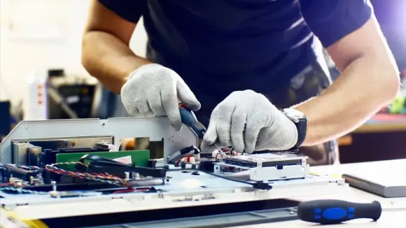 Manos con guantes reparan un componente de computadora con una herramienta, sobre una mesa blanca.