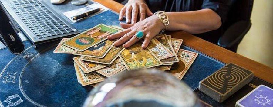 Des mains disposant des cartes de tarot sur une nappe bleue, à côté d’un ordinateur portable et d’étuis de jeu de cartes