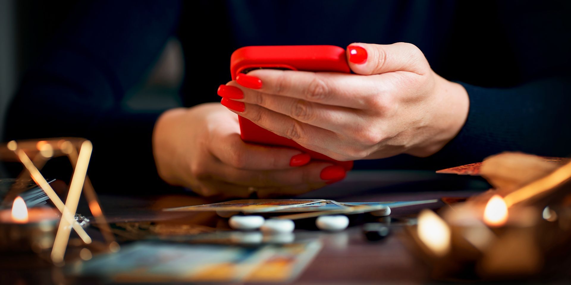 Des mains tenant un smartphone rouge au-dessus d'une table où des pièces de monnaie sont éparpillées et éclairée par une douce lumière de bougie.