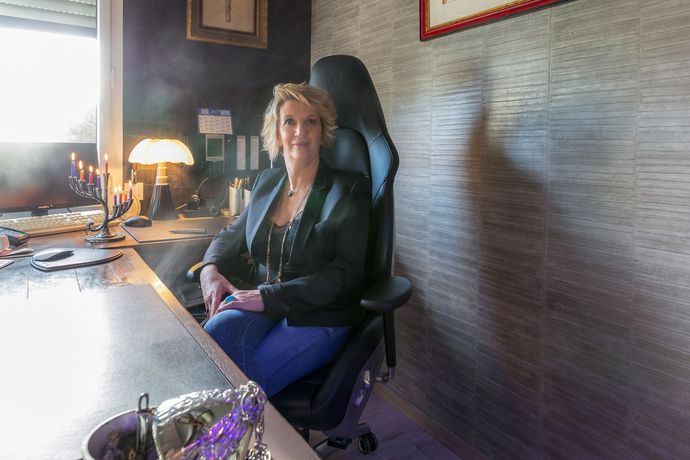 Une personne assise à un bureau, souriante, sur une chaise noire à côté d'une lampe et d'un ordinateur.