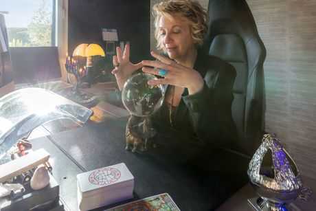 Une femme assise dans un fauteuil noir contemple une boule de cristal posée sur un bureau, entourée de cartes de tarot et de lampes scintillantes.