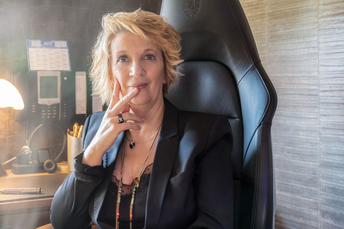 Une femme en blazer noir, assise sur une chaise de bureau, tient un stylo près de ses lèvres, à côté d'une lampe de bureau.