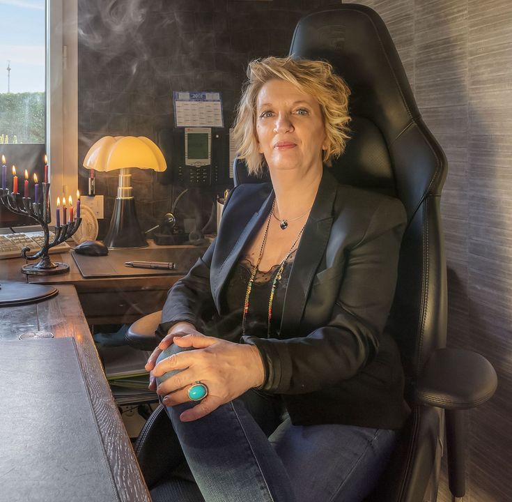 Une femme assise à un bureau dans un bureau sombre, face à la caméra, une lampe allumée à côté d'elle.