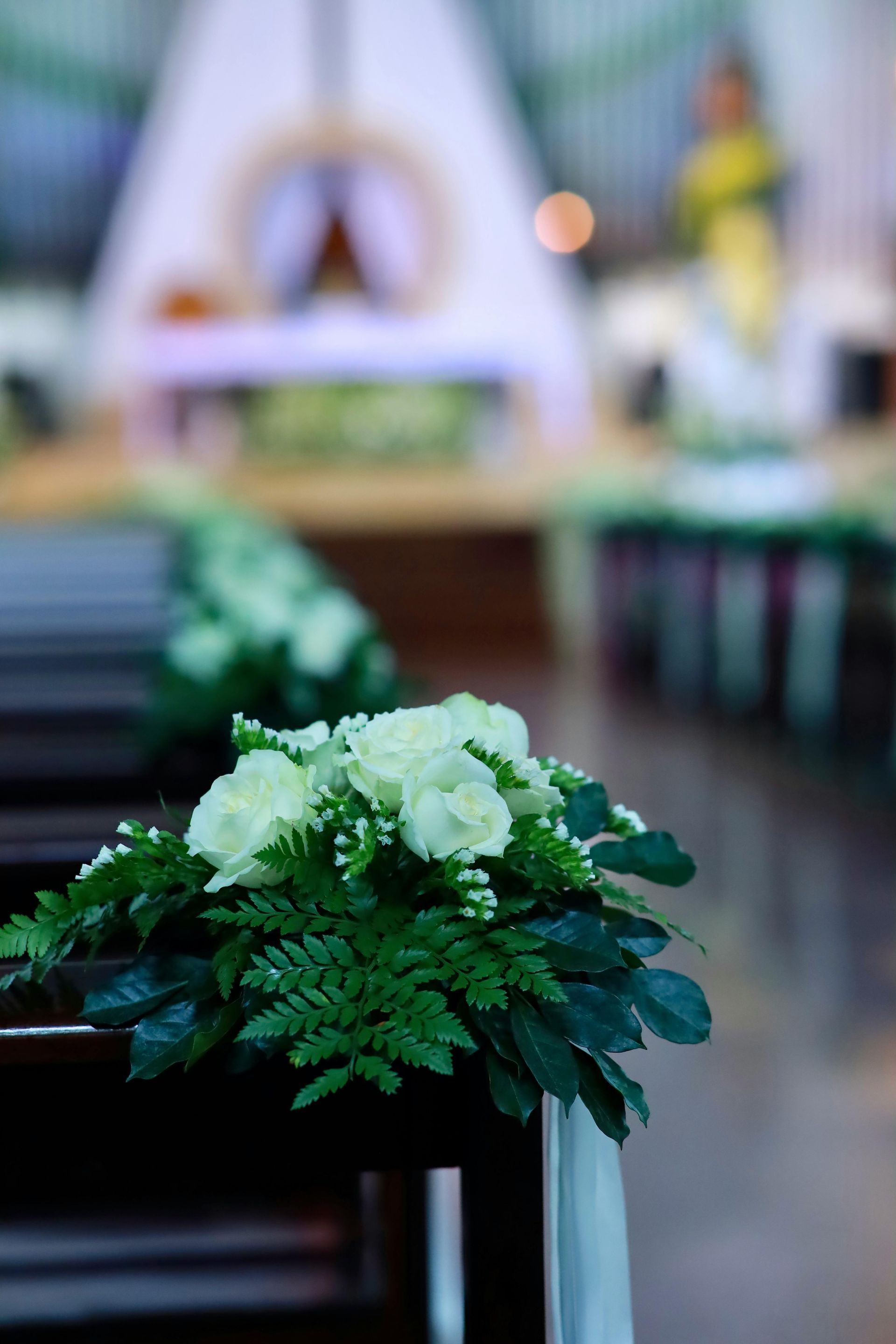 Composition florale sur un banc d'église, avec du feuillage vert et des fleurs blanches au premier plan et un autel flou en arrière-plan.