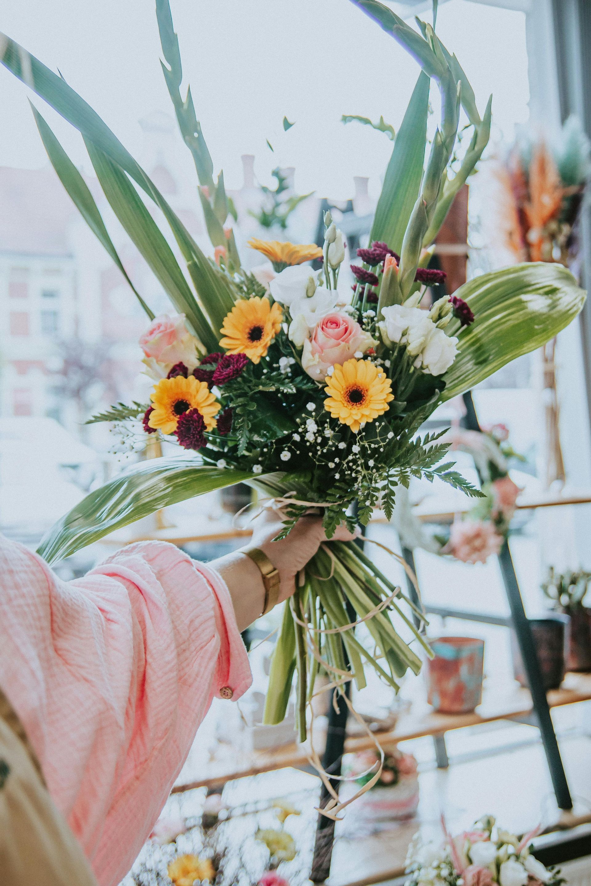 Une main tenant un bouquet coloré composé de fleurs jaunes, de verdure et de fleurs roses dans une pièce lumineuse