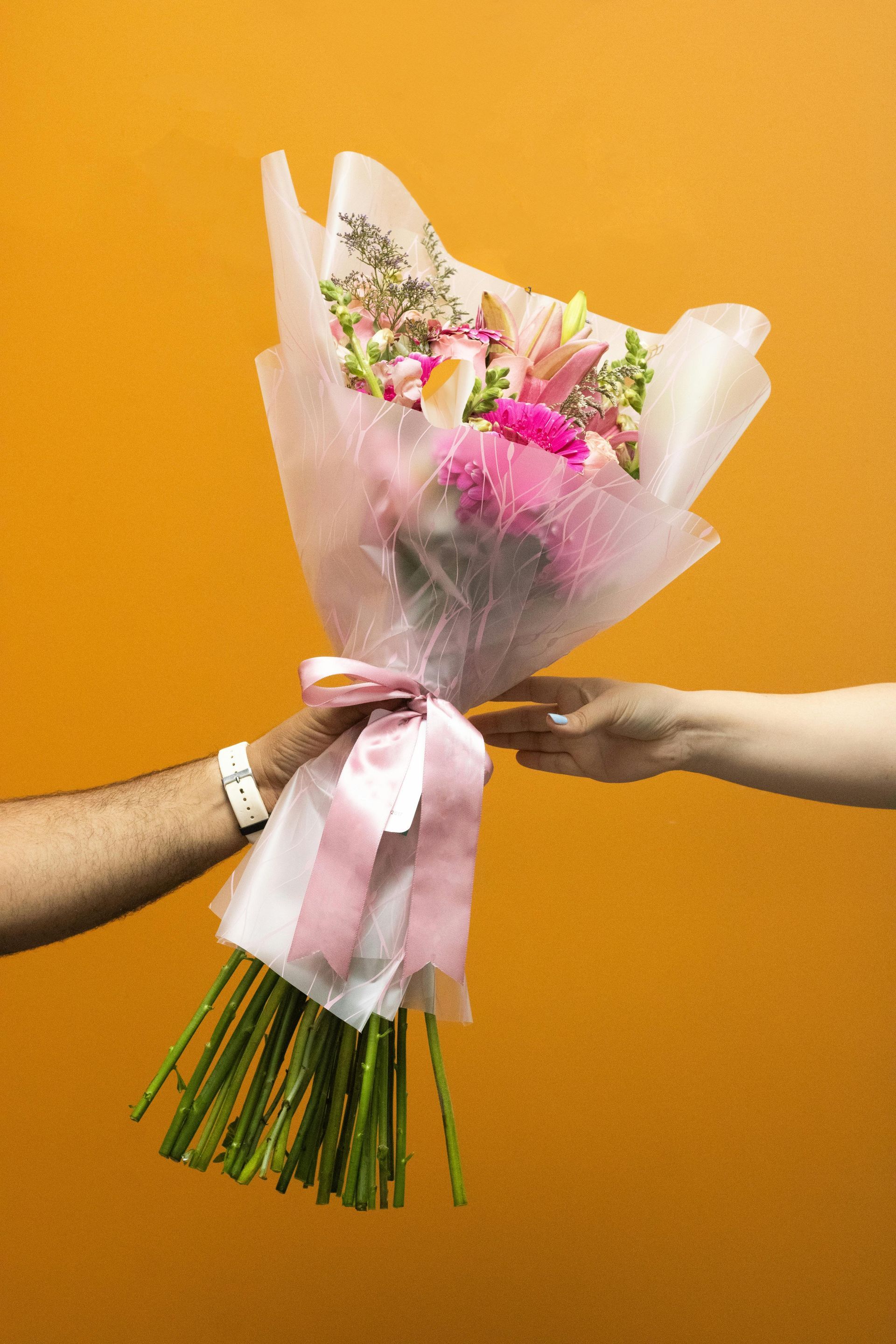 Un bouquet de fleurs roses enveloppé dans du papier rose est échangé entre deux personnes sur fond orange.